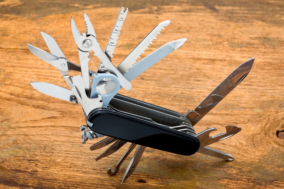 Na zdjęciu znajduje się rozłożony multitool. Jego korpus jest czarny, narzędzia srebrne, a tło ma kolor jasnego drewna.