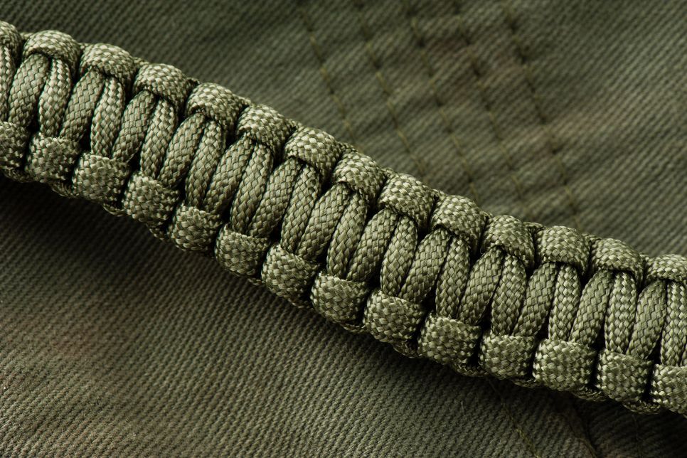 Na zdjęciu widnieje bransoletka z linki paracord w kolorze khaki, ułożona na materiale w tej samej barwie.