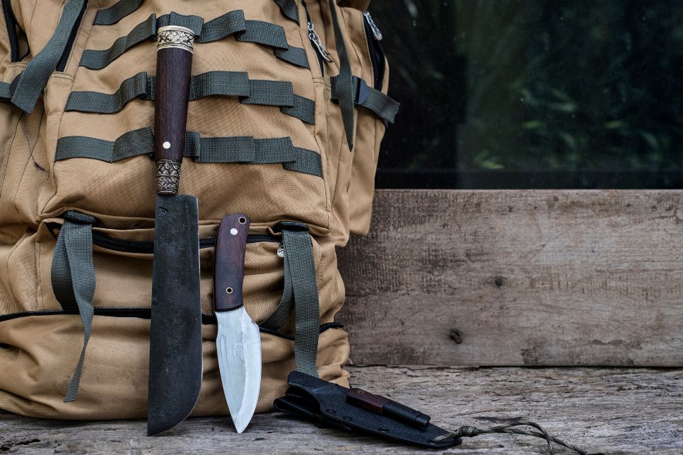 Na zdjęciu znajdują się dwa noże survivalowe i kabura, ustawione na tle plecaka survivalowego w kolorze jasnego brązu.