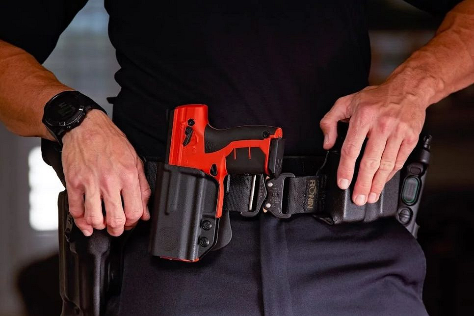 Zdjęcie przedstawia pomarańczowy pistolet na kule gumowe znajdujący się w policyjnej kaburze przypiętej do pasa męzczyzny.