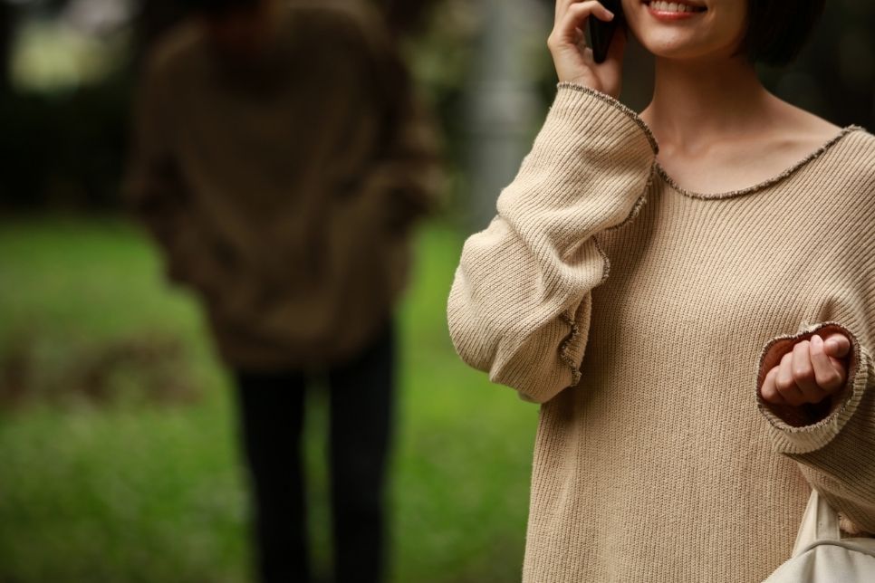 Na zdjęciu widnieje sylwetka kobiety w beżowym swetrze, kt&oacute;ra rozmawia przez telefon. Za nią czai się męzczyzna w czarnym stroju.