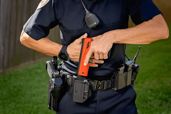 Zdjęcie przedstawia mężczyznę chowającego pomarańczowy pistolet na kule gumowe do policyjnej kabury.